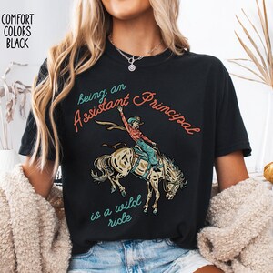 May include: Black t-shirt with the text "Being an Assistant Principal is a wild ride." The graphic features a person riding a bucking horse. The shirt also has the words "Comfort Colors Black" printed on the top left.