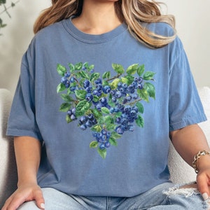May include: A light blue t-shirt with a watercolor design of blueberries in the shape of a heart.