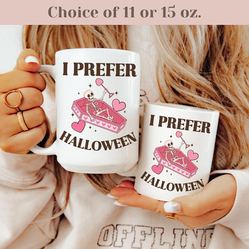 May include: Two white ceramic mugs with a black and pink design. The mugs feature a skeleton in a coffin with hearts and the text "I Prefer Halloween". The mugs are available in 11 or 15 ounce sizes.