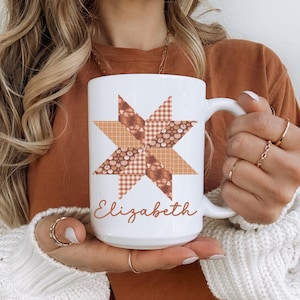 May include: White ceramic mug featuring a patchwork star design in shades of brown, orange, and cream. The name "Elizabeth" is written in a cursive font below the star. The mug has a large handle.