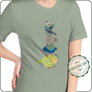 Puede incluir: Una camiseta verde con una pila de pájaros coloridos y un pájaro verde y blanco sobre un fondo verde. Los pájaros están etiquetados con sus nombres en texto negro. El texto "DESIGNED BY ARTIST ORIGINAL ART DESIGNED BY ARTIST" está en un círculo en el lado derecho de la camiseta.