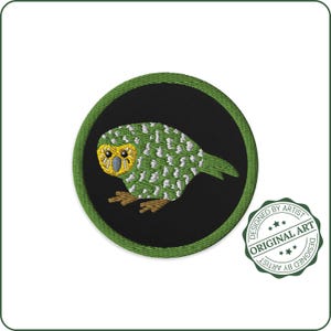 Peut inclure: Patch brodé représentant un oiseau vert et blanc aux yeux jaunes sur fond noir. Le patch est entouré d'une bordure verte et comporte un cercle blanc avec le texte "ORIGINAL ART" en noir.