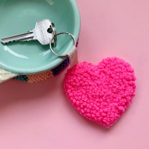 May include: A pink heart-shaped keyring with a silver key and key ring attached. The keyring is made of a fuzzy material and is sitting on a light pink surface.