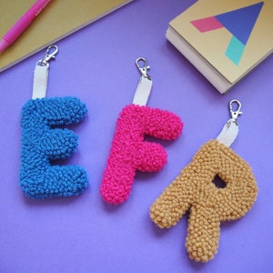 May include: Three colorful, textured letter keychains: a blue "E", a pink "F", and a tan "P". Each letter is attached to a white strap and silver clasp. The keychains are arranged on a purple surface, with a notebook and pen in the background.