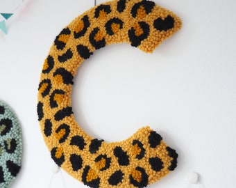 Puede incluir: Una gran letra C amarilla y negra hecha de hilo con un patrón de estampado de leopardo. La letra está colgada en una pared blanca.