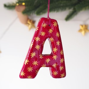 May include: A glossy red letter A ornament featuring a repeating pattern of small gold star-shaped motifs. The letter is suspended by a thin pink string attached to the top, creating a festive hanging decoration with a smooth, reflective finish.