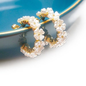 Puede incluir: Un par de pendientes de aro dorados adornados con pequeñas cuentas blancas en forma de flor. Los pendientes se colocan sobre un cuenco de color turquesa con un borde dorado. Joyería delicada y femenina.