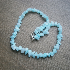Op de afbeelding: Een lichtblauwe edelsteenketting gemaakt van kleine, onregelmatig gevormde stenen. De ketting is geregen aan een zilveren ketting en heeft een zilveren sluiting.