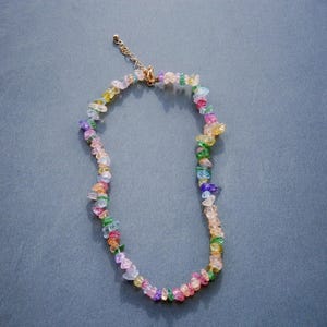 May include: A multi-coloured necklace with small, irregularly shaped beads in pink, purple, green, yellow, and blue. The necklace has a gold-coloured chain and clasp. The beads are strung together to form a choker-style necklace.