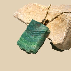 May include: A green and blue stone pendant necklace with a gold-coloured chain. The stone has a rough, natural texture and is set against a beige rock. The necklace is a unique piece of jewellery.