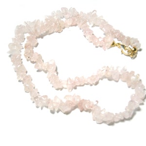 May include: A necklace made of small, irregular rose quartz stones. The pale pink stones are strung together, forming a necklace with a gold-coloured clasp. The necklace is displayed against a white background.