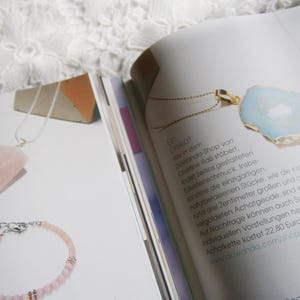 May include: A close-up of a light blue agate pendant necklace with a gold chain. The pendant is on a white surface with text about the necklace and the shop it is from. The text reads "Unikat der in dem DaWanda-Shop von Christine Ra&szlig; st&ouml;bert, indef zeitlos gestalteten Edelsteinschmuck. Insbe sondere die einzigartigen, naturbelassenen St&uuml;cke, wie die Katte mit der rund drei Zentimeter gro&szlig;en und an den R&ouml;nderm vergoldeten Achatgeode, sind echte Eyecatcher Auf Nachfrage k&ouml;nnen auch Schmuckst&uuml;cke nom individuellen Vorstellungen hergestel werden. Achatkette kostet 22,80 Euro. www.dawanda.com/shop/Deramick"