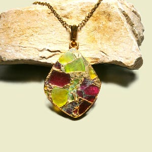 Puede incluir: Un collar dorado con un colgante que presenta un mosaico de piedras de colores de forma irregular. Los colores del colgante incluyen tonos de verde, rojo y amarillo, engastados en un marco dorado. El collar se exhibe sobre una piedra clara.