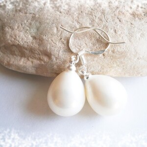 May include: A pair of pear-shaped, off-white earrings with silver-colored hooks. The earrings are displayed on a light-colored, textured stone against a white background. The earrings are a simple, elegant design.