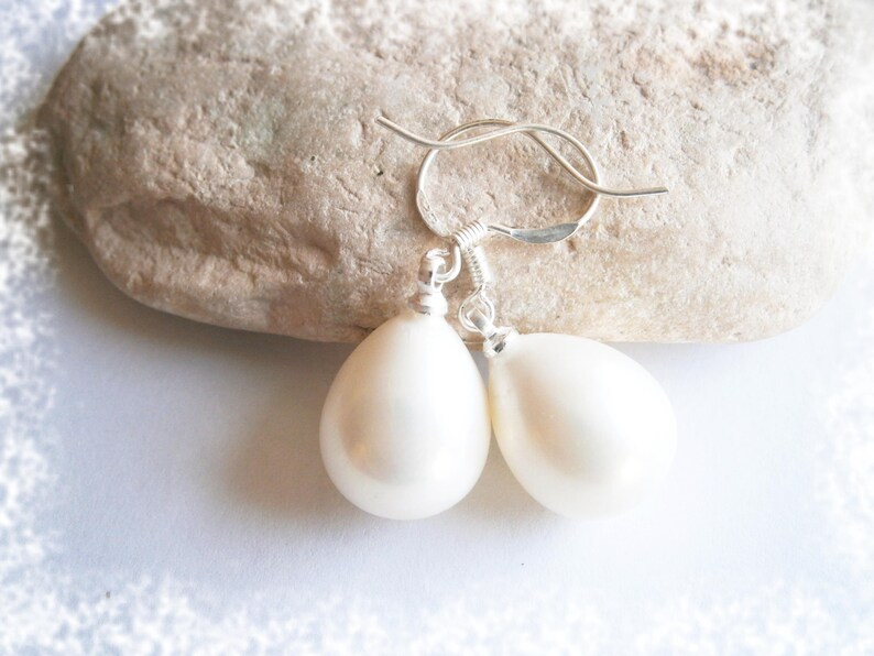 May include: A pair of pear-shaped, off-white earrings with silver-colored hooks. The earrings are displayed on a light-colored, textured stone against a white background. The earrings are a simple, elegant design.