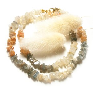 May include: A strand of gemstone beads in shades of grey, white, and orange. The beads are irregular in shape and have a natural, rough texture. The beads are strung on a clear cord and are accented with a small gold clasp.