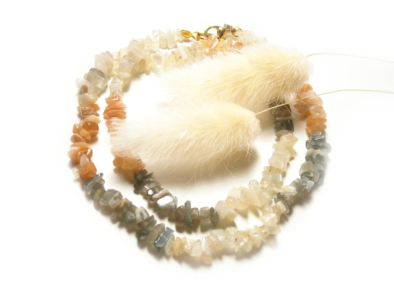 May include: A strand of gemstone beads in shades of grey, white, and orange. The beads are irregular in shape and have a natural, rough texture. The beads are strung on a clear cord and are accented with a small gold clasp.