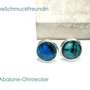 Könnte beinhalten: Ein Paar runde Ohrstecker mit schillernden blauen und grünen Abalone-Muscheleinlagen in silberfarbenen Metallfassungen. Der Text "DeineSchmuckFreundin" und "Abalone-Ohrstecker" ist im Bild zu sehen, zusammen mit einem Betonblock.