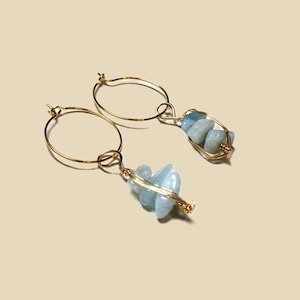 Könnte beinhalten: Ein Paar goldene Creolen mit hellblauen Aquamarinsteinen. Die Steine sind in einem Drahtbüschel gewickelt und an den Reifen befestigt. Die Ohrringe werden vor einem neutralen Hintergrund präsentiert.