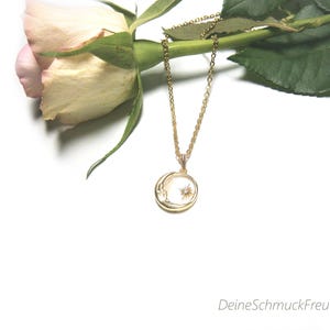 Puede incluir: Un collar de tono dorado con un colgante circular que presenta un diseño de media luna y una pequeña estrella. El colgante cuelga de una delicada cadena dorada. El texto "DeineSchmuckFreundin" es visible en la esquina inferior derecha.