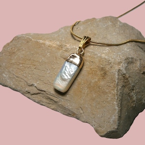 Könnte beinhalten: Halskette mit rechteckigem Anhänger und goldfarbener Kette. Der Anhänger hat eine goldfarbene Kappe und einen weißen, perlmuttartigen Körper. Die Halskette wird auf einem rauen, beigefarbenen Stein vor rosa Hintergrund präsentiert.