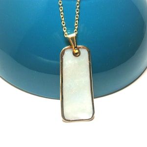 Puede incluir: Collar con cadena dorada y colgante rectangular. El colgante tiene un marco dorado y un centro rectangular de color claro, posiblemente iridiscente. El fondo es azul verdoso borroso.