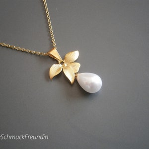 Puede incluir: Un collar dorado con un colgante floral y una perla blanca en forma de gota. El collar tiene una cadena delicada. El colgante presenta un diseño texturizado y detallado. El fondo es gris.