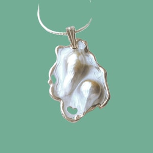 Puede incluir: Un collar con un colgante de plata con una perla barroca blanca y dorada engastada en un dise&ntilde;o de concha.