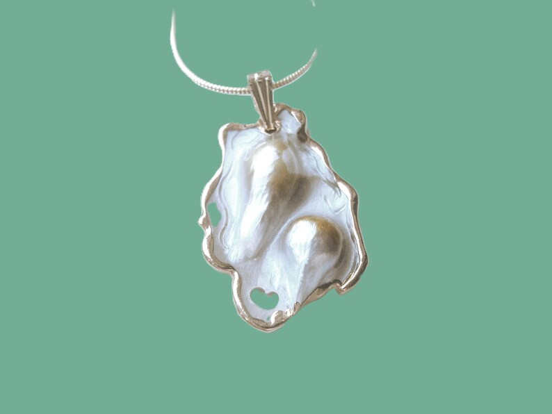Puede incluir: Un collar con un colgante de plata con una perla barroca blanca y dorada engastada en un dise&ntilde;o de concha.