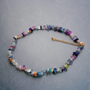 May include: A multi-coloured gemstone necklace. The necklace is made of small, irregularly shaped stones in shades of purple, blue, green, orange and clear. It is strung on a delicate gold-coloured chain, creating a vibrant accessory.
