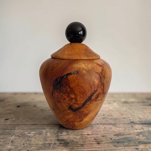 May include: A handcrafted wooden vessel with a natural light brown grain and dark swirling patterns. The container features a removable lid topped with a smooth, polished black spherical finial, creating a contrast between the wood and the orb.