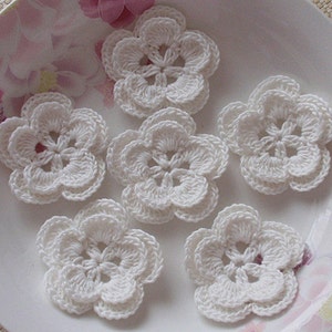 May include: Six white crocheted flower appliques on a white plate. Each flower features five petals and a detailed centre. The flowers are made from soft, textured yarn, ideal for craft projects.