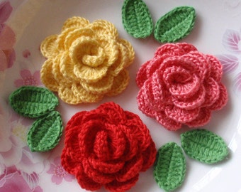 Puede incluir: Tres flores de ganchillo en amarillo, rosa y rojo con hojas de ganchillo verdes. Las flores están dispuestas en un plato blanco con un patrón floral.