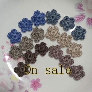May include: Assortment of crocheted flower embellishments in shades of blue, brown, and beige. The flowers are arranged in a semi-circle on a white surface. The text "On sale" is written in yellow letters on the surface.