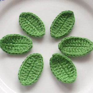May include: Six small, handmade green crochet leaves arranged in a circular pattern. Each leaf has a detailed, textured surface, showcasing the individual stitches. The leaves are a vibrant green color, suggesting a natural theme.