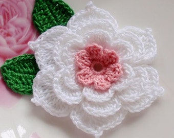May include: A handmade crochet flower with white petals and a pink center. Two green crochet leaves are positioned to the left of the flower. The flower is a craft item, possibly for embellishment or decoration.