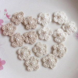 May include: A collection of 12 crocheted flower embellishments in cream and beige colors. Each flower has a pearl bead in the center.