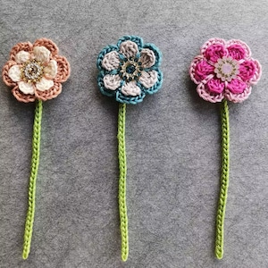 May include: Three crocheted flowers with green stems. The flowers are pink, blue and white, and each has a sparkly center.