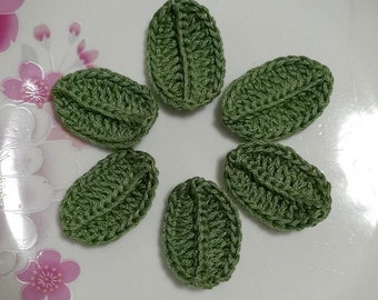 May include: Six green crocheted leaf shapes arranged in a circle on a white surface with pink floral designs.