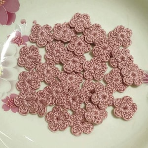 May include: A collection of small, crocheted flowers in a dusty rose color. The flowers are arranged on a white surface, showcasing their delicate, five-petal design. These handmade floral embellishments are ideal for various craft projects.