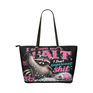 May include: A black tote bag featuring a graphic of a raccoon wearing pink sunglasses, surrounded by salt, pink flowers, and the text "I season with SALT I Don't sugarcoat shit" with a salt shaker labeled "EXTRA SALTY" at the bottom right corner.