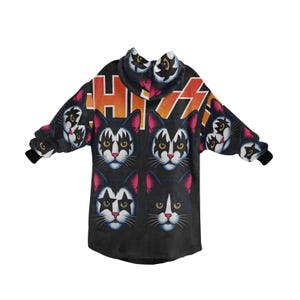 Puede incluir: Manta con capucha negra con un patr&oacute;n repetido de caras de gato maquilladas, que recuerdan a la banda KISS. La capucha y las mangas muestran el logotipo de la banda en naranja y blanco. La manta tiene pu&ntilde;os negros.