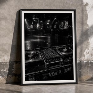 May include: A black and white framed wall art print depicting a DJ booth with two turntables and a central mixer. The scene overlooks a dark, empty nightclub dance floor featuring a disco ball hanging from the ceiling and dim ambient lighting.