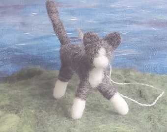 May include: A handmade felt cat figurine in gray and white, standing on a green surface with a blue and white painted background. The cat has a white chest, paws, and face, with its tail raised.