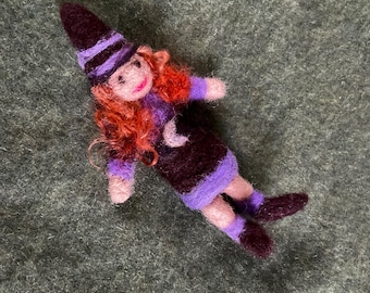May include: A small felt doll wearing a purple and black striped hat, a purple and brown dress with a crescent moon design, and purple and brown shoes. The doll has red hair and a smiling face.