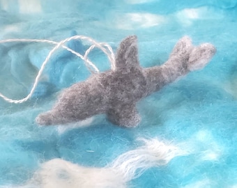 May include: A gray felted dolphin ornament with a white string for hanging. The dolphin has a dorsal fin and a tail fin. The background is a mix of blue and white fibers, creating a sea-like effect.