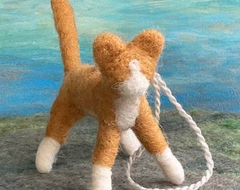 May include: A hand-felted orange and white cat with a white tail, standing on a gray surface with a white string attached to its neck.