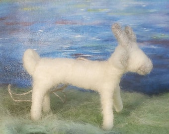 May include: A white felt animal figurine resembling a dog or a lamb, standing on a green and blue fuzzy surface. The figurine has gray ears and a long tail.