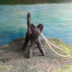 May include: A grey felt cat with a white string attached to its back. The cat is standing on a grey and green surface.