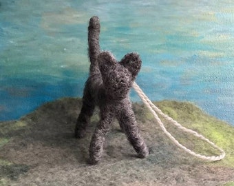 May include: A gray felt cat with a white string attached to its back. The cat is standing on a gray and green surface.